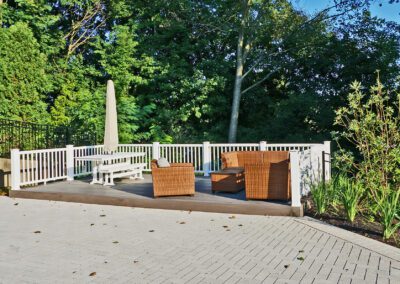 A paved patio area with a white railing, featuring outdoor wicker furniture and surrounded by greenery.
